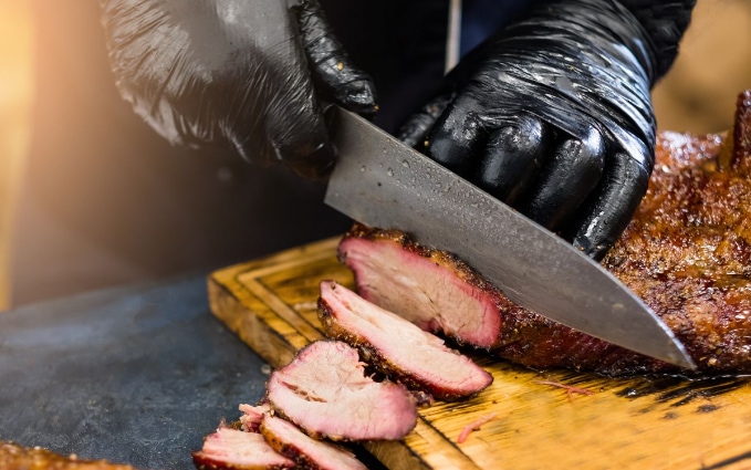 Close-up of gloved hands slicing smoked brisket with a large knife.