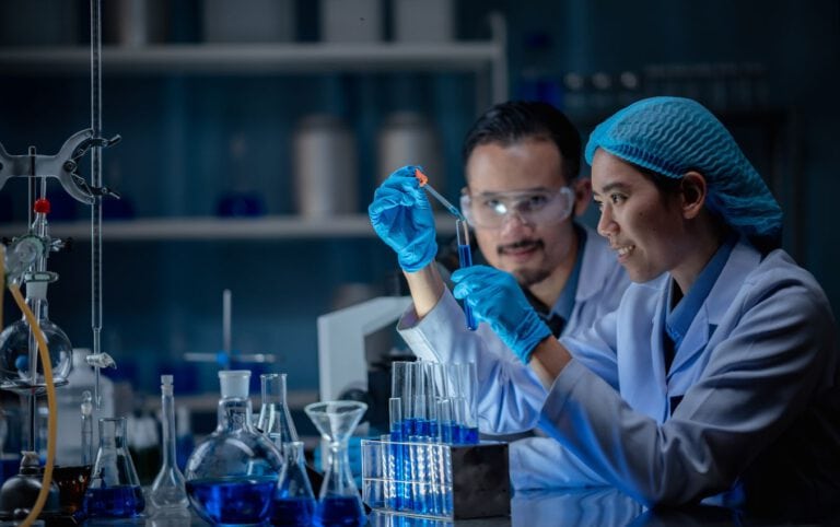 Two scientists in lab coats and gloves conduct experiments, using pipettes and test tubes.